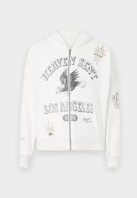 Felpa bianca con zip, decorata con la scritta grafica "HEAVEN SENT LOS ANGELES 1971", un design di un'aquila e toppe decorative tra cui un sole e un mazzo di carte.