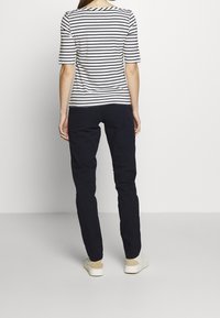 Pruhované tričko s krátkým rukávem v navy a bílé; padnoucí kalhoty v tmavě navy; bílé boty; viděno zepředu na jednobarevném pozadí.