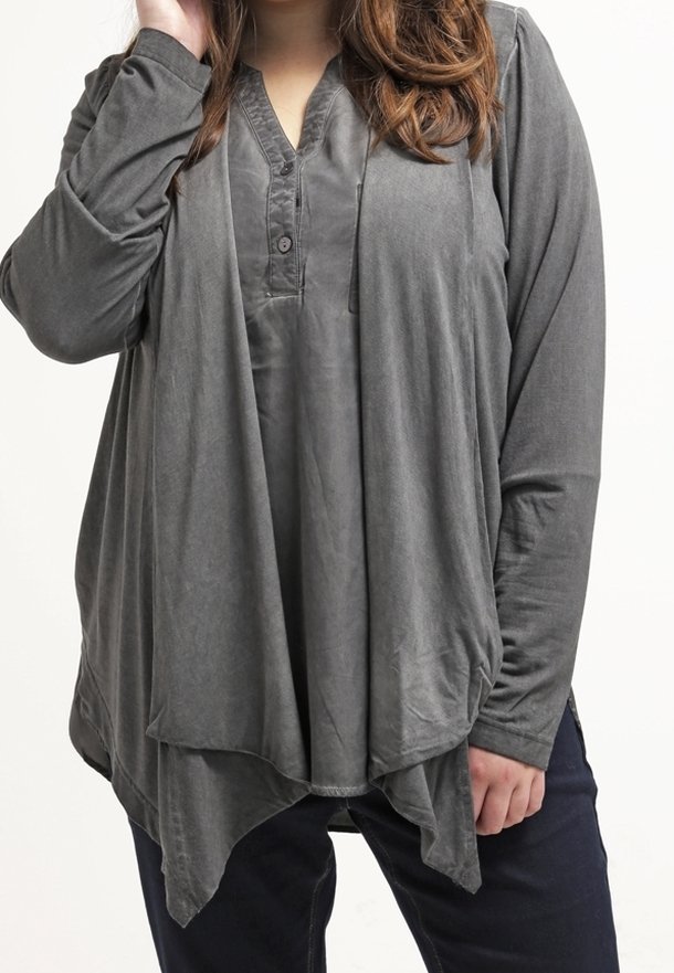 Femme portant un cardigan gris ample à manches longues drapé sur une chemise grise boutonnée, associé à un pantalon foncé, debout devant un fond blanc.