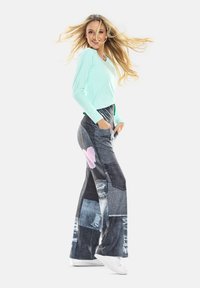 Mintgrünes Langarmshirt kombiniert mit Jeans in Patchwork-Optik und weitem Bein, das ausgefranste Kanten und ein rosa Herz-Akzent aufweist, getragen mit weißen Sneakers.