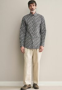 Langärmliges, knöpfbares Hemd mit floralem Muster auf dunklem Hintergrund, kombiniert mit cremefarbenen Hosen und braunen Schuhen.