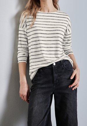 Femme portant une chemise blanche à manches longues avec des rayures horizontales grises et un jean noir taille haute, debout contre un mur gris.