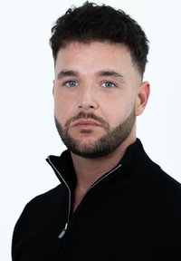 Close-up van een man die een zwart zip-up sweater draagt met een hoge kraag en zilveren ritsdetails. Gladde huid en kort, donker haar.