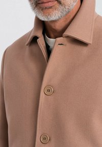 Abrigo de lana beige con cuello tipo solapa, que presenta tres botones marrones y una textura suave, con primer plano que detalla la suavidad de la tela.