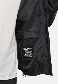 Veste noire imperméable avec un intérieur lisse, arborant une étiquette TEXAPORE. Fermeture éclair à l'avant et poche intérieure pour le rangement.