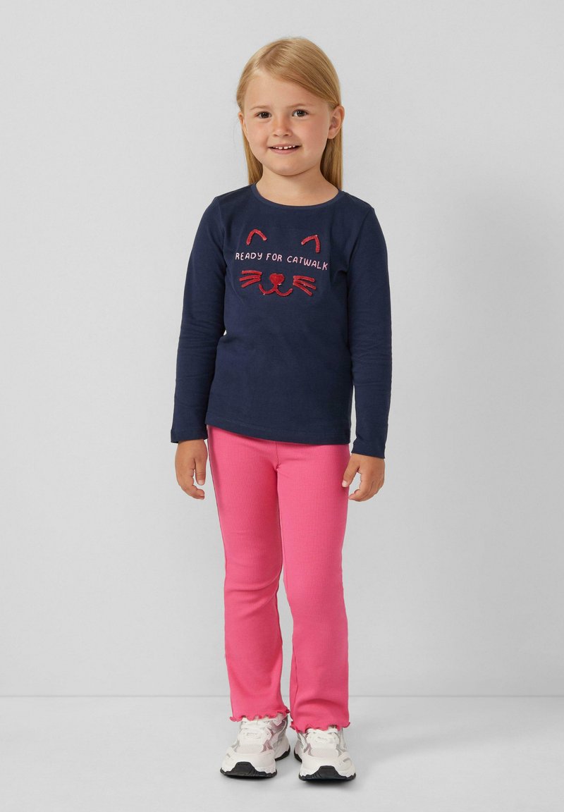 Marineblaues Langarmshirt mit Katzengesichtdesign und rotem Text; rosa Rippleggings; Kind steht in weißen Turnschuhen vor einem schlichten Hintergrund.