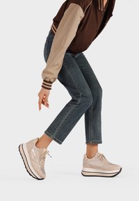 Sneakers piattaforma beige con finitura lucida, tomaia in tessuto e sintetico, lacci testurizzati e suola nera, indossati con jeans alla caviglia.