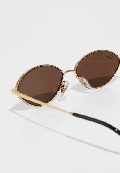 Lunettes de soleil en métal doré avec des verres marron. Forme ovale, dotées de coussinets de nez et d'une embout en plastique noir sur les branches. Monture légère avec une finition brillante.