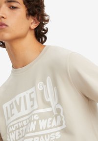 Camiseta de manga corta beige con estampado gráfico en blanco que presenta "Levi's" y un diseño de cactus. Textura suave con cuello redondo.