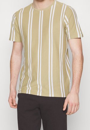 Mand iført beige T-shirt med korte ærmer med lodrette hvide striber og sorte bukser, vist fra nakke til hofter mod en ensartet baggrund.