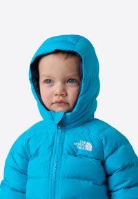 Siniste silmadega väikelaps, kes kanab ere sinist North Face kapuutsiga talvejope, vaadates kergelt kõrvale ühevärvilise tausta ees.