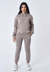 Grijze katoenen hoodie met een kangoeroezak en wit logo, gecombineerd met bijpassende tapersweatpants en witte sportschoenen.