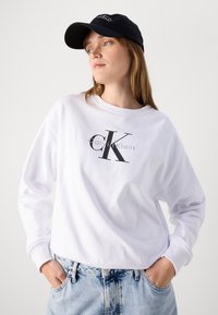 Sweatshirt blanc avec un grand logo noir "CK" et le texte "calvinKlein" ; associé à un jean en denim bleu et une casquette noire.