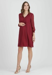 Těhotenské šaty z burgundské chiffon látky s dlouhými rukávy, véčkovým výstřihem, volánkovými detaily a empírovým pasem. Vytvořeno s zlatými sandály na vysokém podpatku.