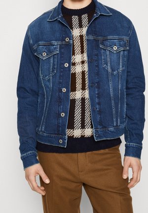 Person, die eine blaue Jeansjacke über einem braun-weiß karierten Pullover trägt, kombiniert mit braunen Hosen, vor einem einfarbigen Hintergrund steht.