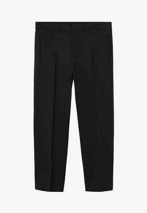 Hombre con camisa negra de manga corta abotonada, pantalones negros a medida y sandalias negras con dos tiras, manos en los bolsillos, fondo beige.