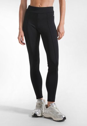 Kvinde iført sorte tætsiddende leggings og sølvfarvede træningssko, stående på en lys baggrund med bar mave synlig.