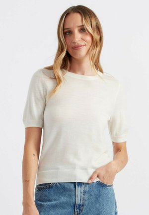 Pull crème à manches courtes avec un col rond, des poignets et un ourlet côtelés, fabriqué dans un tissu tricoté léger. Associé à un short en denim bleu.