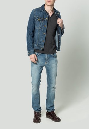 Mann in einer blauen Jeansjacke, schwarzem Henley-Shirt, hellblauen Jeans und dunkelbraunen Schnürstiefeln, der vor einem einfarbigen Hintergrund steht.