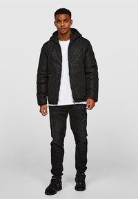 Czarna pikowana kurtka puchowa z kapturem, ozdobiona geometrycznym wzorem. Noszona na białej koszulce i zestawiona z postrzępionymi czarnymi dżinsami.