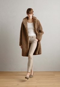 Brun, fluffy overdimensjonert kåpe med stor krage, kombinert med kremfarget topp og lysebeige tettsittende bukser. Fottøyet har en lav hæl.