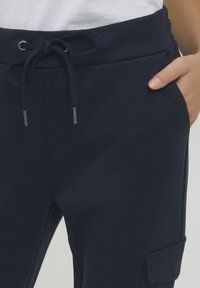 Tmavě modré tepláky vyrobené z měkkého materiálu, s elastickým pasem a šňůrkou a boční kapsou pro uložení.
