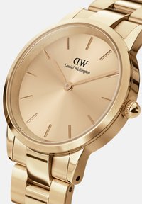 Arany rozsdamentes acél óra fényes felülettel, minimalista számlappal, diszkrét órakészítőkkel és koronával a 3 órás pozícióban. Márka: Daniel Wellington.