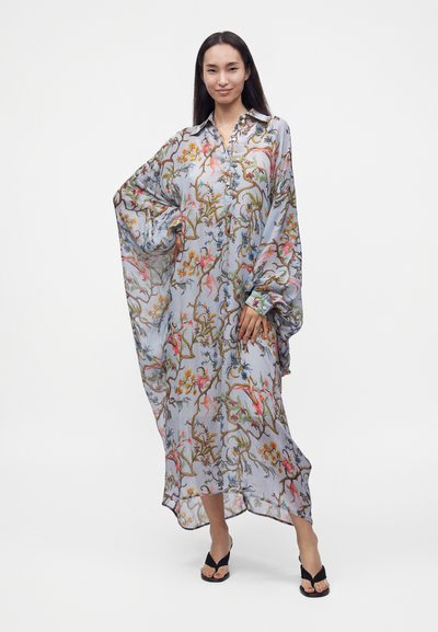 Femme aux longs cheveux foncés portant une longue robe ample bleu clair à motifs floraux et des talons noirs à bout ouvert, debout sur un fond blanc.