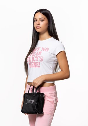 Schwarze Wildleder-Handtasche mit strukturiertem Design, mit glänzendem silbernem Schriftzug "Juicy." Das Model trägt ein weißes Crop-Shirt und pinke Jogginghosen.