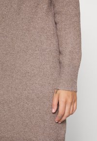 Maglione di lana marrone con polsini a coste e una texture morbida. Mano parzialmente visibile, con tatuaggi contro il tessuto.