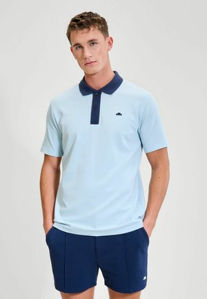 Joven con camiseta tipo polo azul claro con cuello azul marino y pantalones cortos azul marino, de pie con las manos en los bolsillos contra un fondo liso.