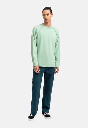 Mintgroene lange mouwen pullover met raglanmouwen, gecombineerd met donkerteal kleurige broek met kniestukken en zwarte sneakers.