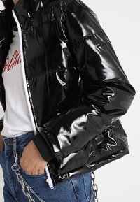 Czarna błyszcząca kurtka puchowa z zamkiem z przodu i ściągaczami na rękawach, o gładkiej, refleksyjnej teksturze i pikowanym wzorze.