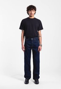 Jeans in denim blu scuro con design a gamba dritta, indossati con una t-shirt nera aderente. Il modello è in piedi con scarpe nere, su sfondo neutro.