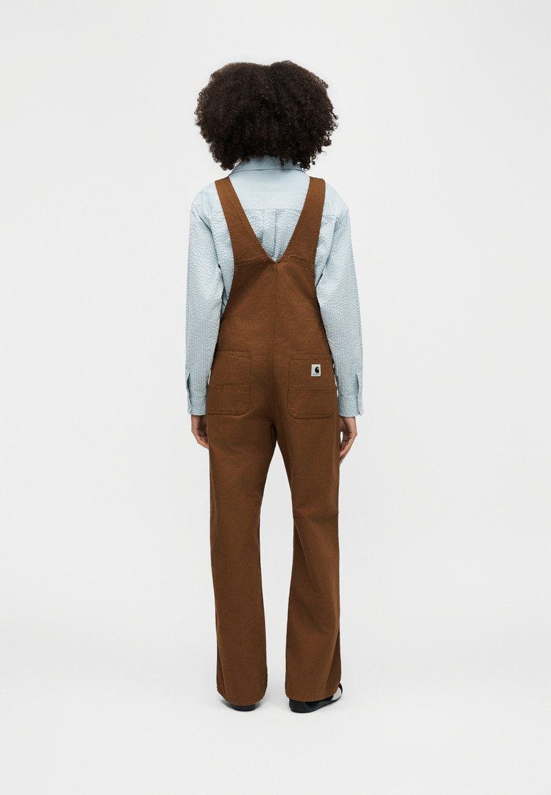 Person with curly hair wearing light blue shirt and brown overalls standing facing away against plain background.