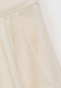 Tissu couleur crème avec des plis cousus et une ceinture, montrant la texture et les détails des coutures sur une surface lisse.
