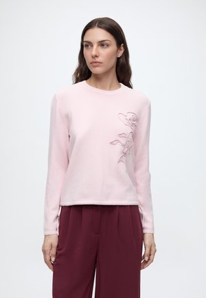 Femme aux longs cheveux bruns porte un pull rose clair avec broderie florale et un pantalon bordeaux taille haute, debout devant un fond blanc uni.