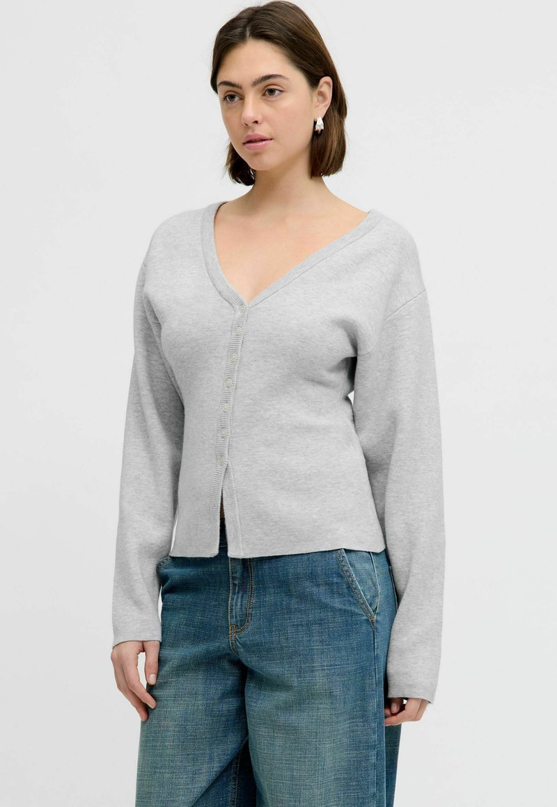 Vrouw draagt een lichtgrijs gebreid vest met knopen, V-hals en lange mouwen, gecombineerd met losse blauwe spijkerbroek, staand tegen een effen achtergrond.