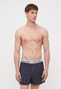 Shorts de bain bleu marine avec une texture douce, taille élastique portant une impression de logo, et coupe ample. Le modèle se tient torse nu.