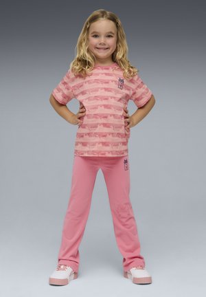 Joven niña de pie con confianza, con las manos en las caderas, vistiendo una camisa de rayas rosas, pantalones a juego y zapatillas blancas con rosa sobre un fondo gris.