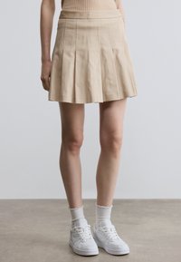 Beige geplooide rok met een gladde textuur, nauwsluitende tailleband en uitlopende zoom, gecombineerd met witte sneakers en gestructureerde enkelsokken.