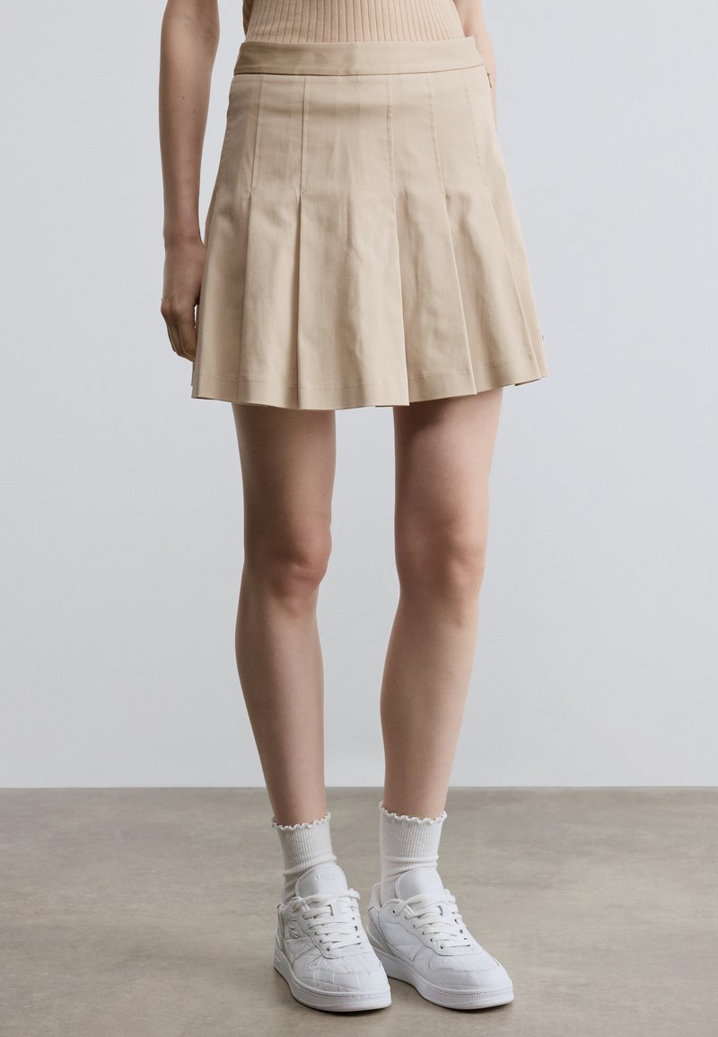 Beige geplooide rok met een gladde textuur, nauwsluitende tailleband en uitlopende zoom, gecombineerd met witte sneakers en gestructureerde enkelsokken.