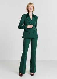 Blazer verde doppiopetto con colletto in satin, abbinato a pantaloni svasati coordinati. La texture liscia e la vestibilità su misura migliorano il design formale.
