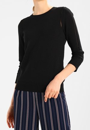 Kvinde iført en sort strikket sweater med kvartærmer og marineblå bukser med hvide og røde lodrette striber.
