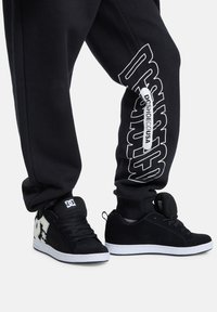 Schwarze Sneaker mit weißen Akzenten und Logodetails, kombiniert mit schwarzen Jogginghosen, die einen weißen, seitlich aufgedruckten Markennamen tragen.