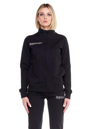 Une femme présente une veste noire à fermeture éclair et un pantalon assorti sur fond blanc.