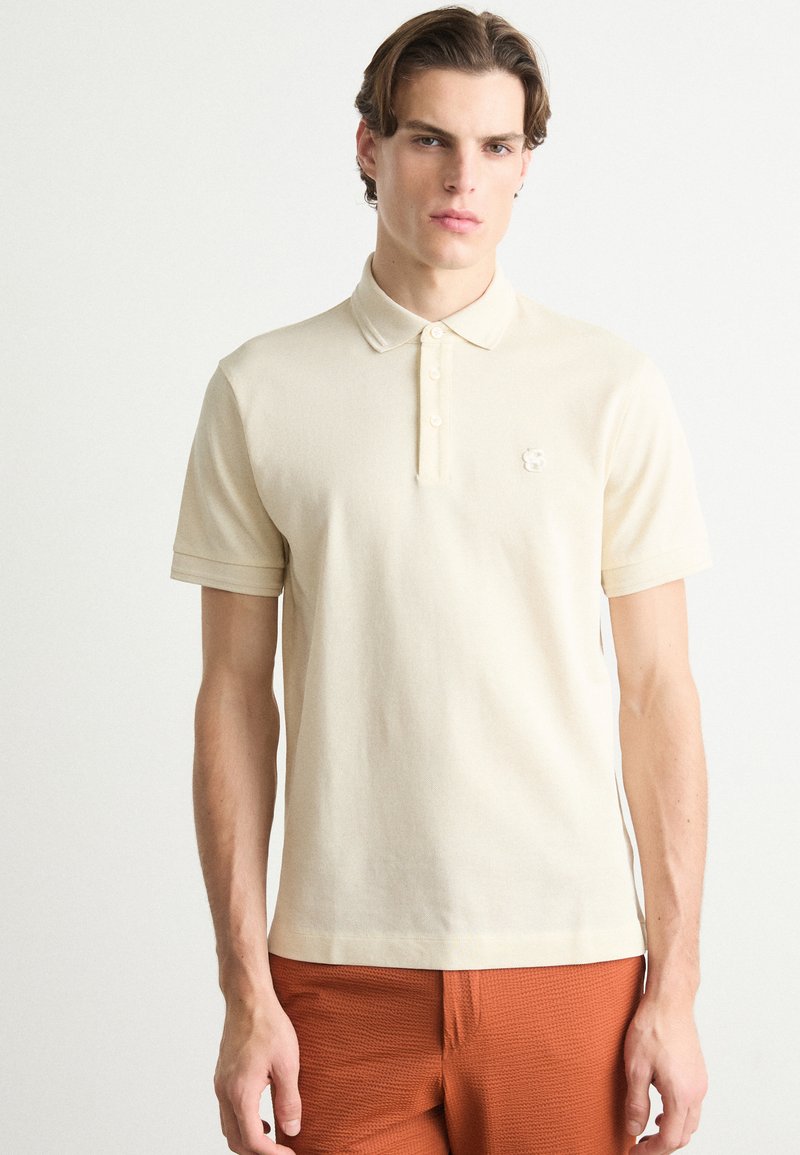 Beige polo skjorte med strukturert overflate, med krage, tre knapper og en liten brodert logo på brystet.