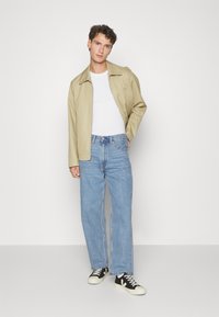 Giacca leggera beige sopra una t-shirt bianca, abbinata a jeans dritti blu chiaro e sneakers nere con suole e dettagli bianchi.