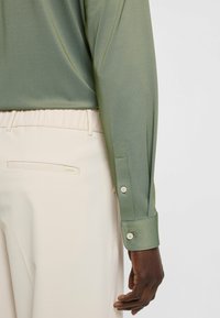 Olijfgroene, lange mouwen overhemd met een gladde textuur en knoopmanchetten, gecombineerd met beige broek met een achterzak en subtiele branding.