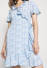 Blå blommig wrapklänning med volangkrage och korta ärmar. Tyget har en ljusblå botten med ett mönster av vita och svarta blad.
